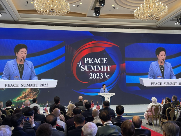 Zwei Frauen stehen auf Podien mit Mikrofonen und sprechen zu einem sitzenden Publikum auf dem Peace Summit in Hong Kong, während im Hintergrund ein Bildschirm mit Text und Logos zu sehen ist.