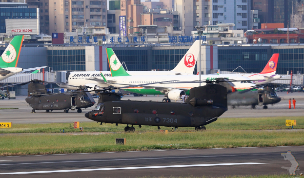 Mehrere Militärhubschrauber auf dem Rollfeld eines Flughafens geparkt, umgeben von Gras und einer Straße, mit Gebäuden, Texttafeln, Pfählen, Fahrzeugen und anderen Gegenständen im Hintergrund und einem Wasserzeichen in der rechten unteren Ecke.