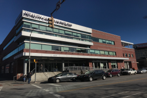 Ein großes Gebäude mit parkenden Autos davor, umgeben von Straßenlaternen, Lampen, Verkehrssignalen, Schildern, Menschen auf dem Gehweg, Geländern, Treppen, Bäumen und einem klaren blauen Himmel, identifiziert als das New York City Health Care Homeless Center.