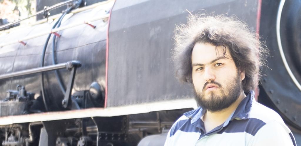 Ein Mann in einem T-Shirt steht neben einem Zug, wobei die Räder und andere Teile des Zugs unten und ein Baum in der oberen linken Ecke zu sehen sind.