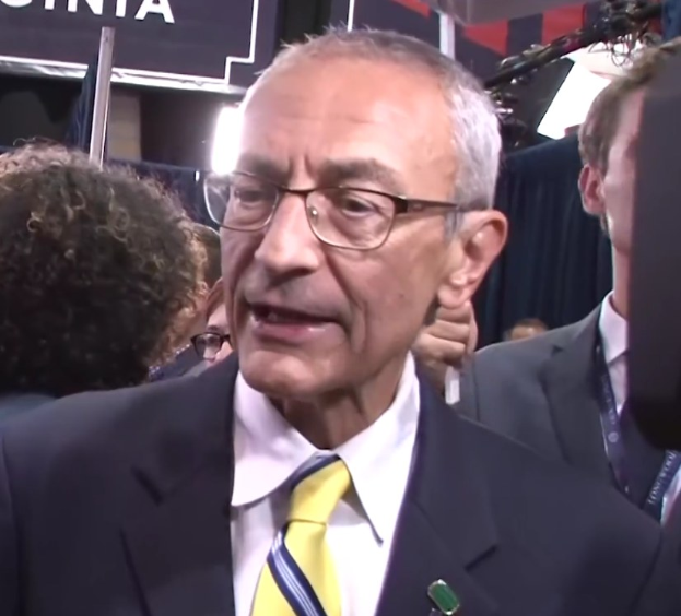 Ein Mann im Anzug und mit Brille spricht zu Reportern, mit einem Mikrofonständer vor ihm und einer Tafel mit Text im Hintergrund.