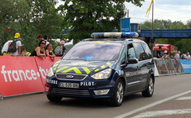Polizeiauto fährt auf einer Straße neben einer Menschenmenge, mit einem Banner auf der linken Seite, Geländern mit Bannern dahinter, Bäumen, einer Brücke, einer Fahne auf einem Mast und einem bewölkten Himmel im Hintergrund.