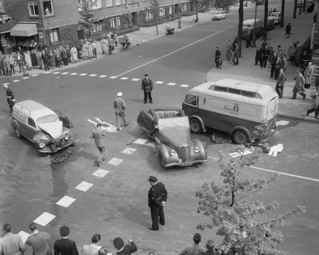 Schwarz-weiß-Szene eines Autounfalls an einer Straßenkreuzung mit mehreren Fahrzeugen, darunter ein Van, und Menschen, die sich darum versammeln; Gebäude, Bäume, Strommasten und Texttafeln sind im Hintergrund zu sehen.