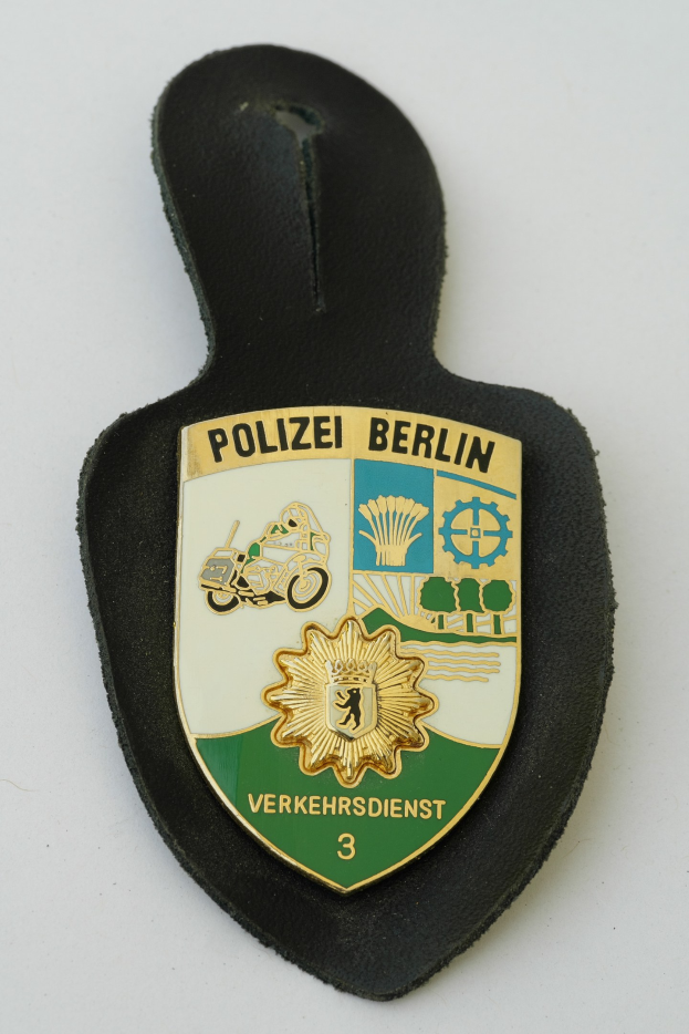 Ein schildförmiges Polizeibadget mit der Aufschrift 'Polizei Berlin' auf einem blauen Hintergrund, einem weißen Stern und einem weißen Rand, das auf einer Oberfläche platziert ist.