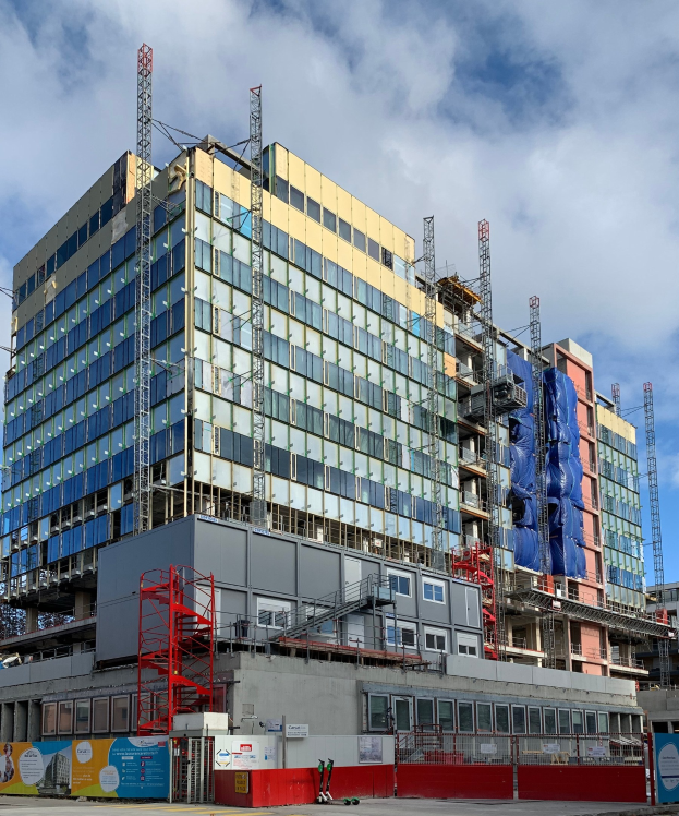 Großes Gebäude im Bau in einer Stadt mit Glasfenstern, Metallrahmen, einer Treppe, einem Zaun, einem Schild, Pfosten, einem Straßenpfosten, Bäumen und einem bewölkten Himmel.