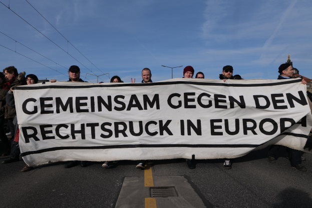 Eine Gruppe von Menschen steht auf der Straße und hält ein Banner mit der Aufschrift "Gemeinsam gegen den Rechtsruck in Europa" in den Händen mit Straßenlaternen, Strommasten, Stromkabeln, Gebäuden und einem bewölkten Himmel im Hintergrund.