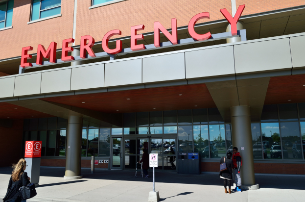 Gruppe von Menschen, die auf ein Notaufnahme-Gebäude mit Glaswänden, Säulen, einem Namensschild und nahen Schildern und einem Mülleimer zugehen.