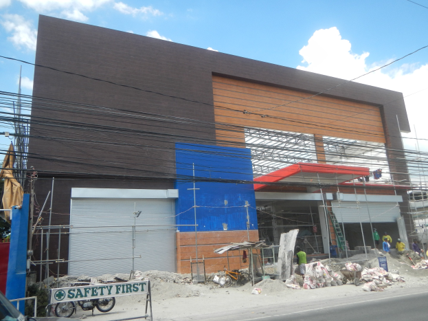 Ein Gebäude im Bau mit einem "Sicherheit geht vor"-Schild, Fahrzeuge auf der Straße, eine Tafel mit Text, einige Menschen in der Nähe und im Hintergrund Bäume, Pfähle, Drähte und der Himmel.