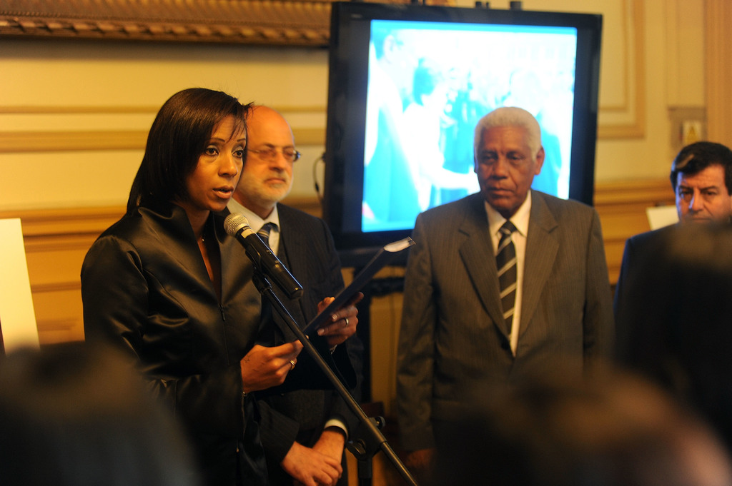 Eine Frau steht an einem Mikrofon auf einer Bühne mit einem Fernseher und einer Wand im Hintergrund.