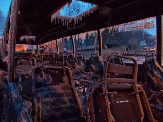 Ein zerstörter Bus mit verkohlten Sitzen und Eiszapfen an der Decke vor dem Hintergrund von Bäumen, Gebäuden und einem klaren Himmel.