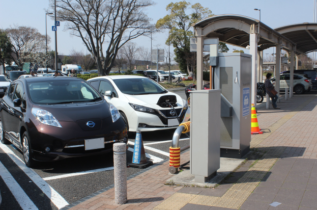 Elektroauto-Ladestation in Japan mit Autos, Verkehrskegeln, einer Person auf dem Gehweg, einem Schuppen, Mästen, Lichtern, Schildern, Bäumen, Pflanzen und einem Himmel im Hintergrund.