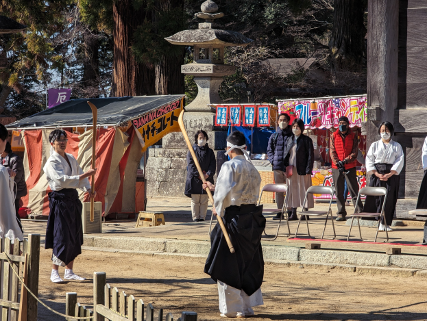 Eine Gruppe von Menschen in traditioneller Kleidung steht im Freien in Kyoto, einige tragen Masken und halten Holzstöcke, während Stühle, Banner, ein Zelt und ein Holzzaun unter einem klaren blauen Himmel zu sehen sind.