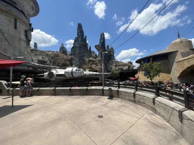 Das Millennium-Falke in Star Wars: Galaxy's Edge mit Besuchern, Schirmen, Zaun, Straßenpfosten, Schild, Bäume, Hügel, Drähte und eine bewölkte Himmel.