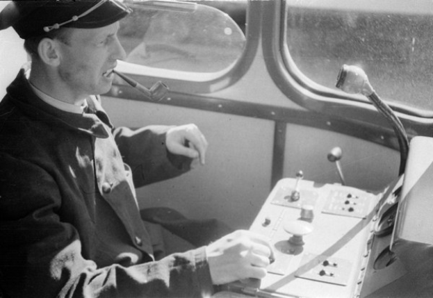 Schwarzes und weißes Bild eines Mannes in einer Mütze und Uniform, der an den Kontrollen eines Zugs sitzt und eine Pfeife raucht, mit sichtbaren Glasfenstern im Hintergrund.