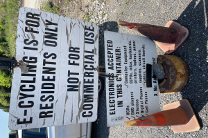 Ein Schild mit der Aufschrift "E-Bike-Fahren ist nur für Bewohner erlaubt" wird an der Seite einer Straße aufgestellt, umgeben von Verkehrskegeln und anderen Gegenständen, mit einem Fahrzeug, Bäumen und einem klaren blauen Himmel im Hintergrund.