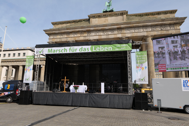 Eine Bühne vor dem Brandenburger Tor in Berlin, Deutschland, mit einem Tisch mit einer Vase, einem Kreuzsymbol, Transparenten mit Text, Lautsprechern und anderen Gegenständen, sowie Fahrzeugen auf der Straße und Gebäuden im Hintergrund.