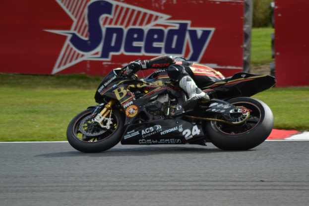 Eine Person in Helm und Schutzkleidung fährt ein Motorrad auf einer Rennstrecke, umgeben von Gras und einer Tafel mit Text im Hintergrund.