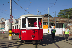 Eine rote und weiße Tram auf einer Stadtstraße mit einigen Passagieren, zwei Personen in grünen Jacken und schwarzen Hosen auf dem Gehweg und verschiedene Gebäude, Fahrzeuge und Bäume im Hintergrund.