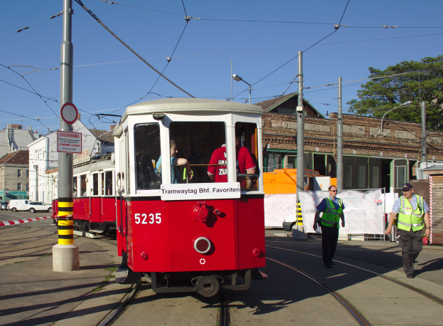 Eine rote und weiße Tram auf einer Stadtstraße mit einigen Passagieren, zwei Personen in grünen Jacken und schwarzen Hosen auf dem Gehweg und verschiedene Gebäude, Fahrzeuge und Bäume im Hintergrund.