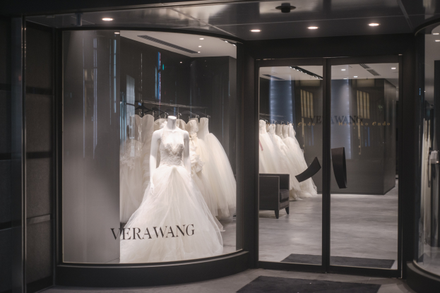 Innenansicht eines Brautmodengeschäfts in Singapur mit Hochzeitskleider auf Schaufensterpuppen und Bügeln, einer Glaswand mit Text, einem Teppich, einem Stuhl und Deckenbeleuchtung.