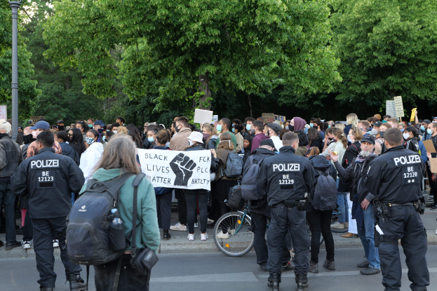 Eine große Gruppe von Menschen nimmt an einer Black Lives Matter Demonstration in Berlin teil, einige halten Schilder, andere tragen Kappen und Taschen, mit einem Fahrrad im Vordergrund und Bäumen und einem Pfahl im Hintergrund.