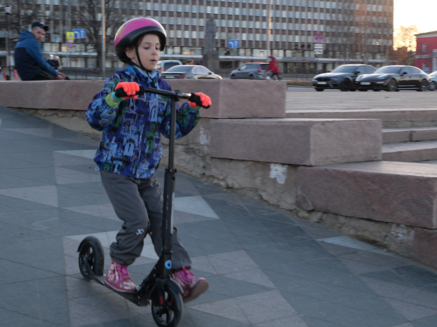 Ein junger Junge in Helm und Handschuhen fährt mit einem Roller eine Treppe auf einem Gehweg hinunter, mit Fahrzeugen, Menschen, Bäumen, Pfählen, Brettern, Gebäuden und einem klaren blauen Himmel im Hintergrund.
