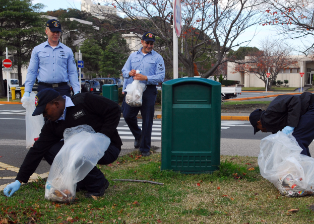 Drei Polizisten in Uniform sammeln Müll an einer Straßenseite, mit Bäumen, Gebäuden, Fahrzeugen und einem Mülleimer im Hintergrund.