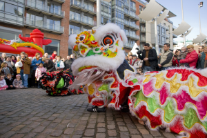 Eine Löwentanzvorstellung während eines chinesischen Neujahrsfestes in Amsterdam, mit einer Zuschauermenge und Menschen, die den Moment mit Kameras festhalten, vor einem Hintergrund aus Gebäuden und einem klaren blauen Himmel.