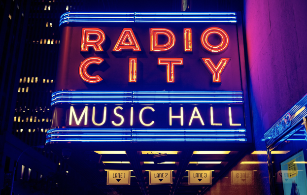 Außenansicht des Radio City Music Hall in New York City bei Nacht, hell erleuchtet und von hohen Gebäuden flankiert, mit einem "Radio City"-Schild und mehreren kleineren Schildern an seiner Basis.