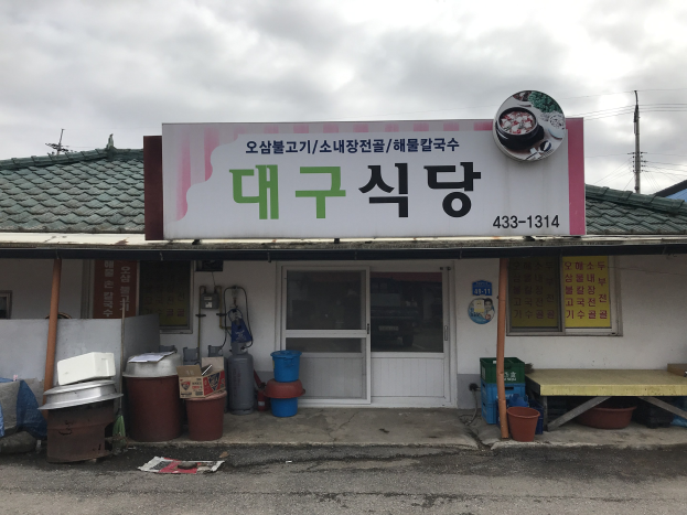 Kleines Gebäude mit einer Tafel, die "Koreanisches Restaurant" lautet, einer Tür, Fenstern, einem Dach, nahegelegenen Behältern, Eimern, einem Tisch, Polen, Drähten, einem anderen Haus und einem bewölkten Himmel.