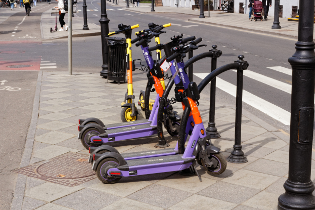 Eine Reihe von E-Scootern, die an einer Straße mit Fußgängern, Radfahrern, einem Müllcontainer im Vordergrund und Gebäuden, Bäumen und Pflanzen im Hintergrund geparkt sind.