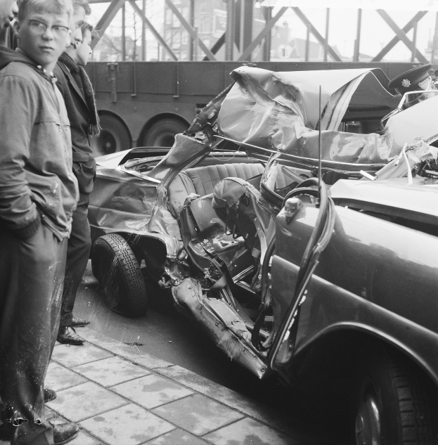 Schwarz-weißes Bild eines verbeulten Autos am Straßenrand mit mehreren Personen in der Nähe, einem Lkw und einer Brücke im Hintergrund.