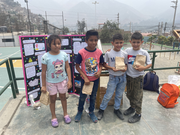 Gruppe von Kindern auf einem Basketballfeld mit Papiertüten in der Hand, mit Vorräten auf dem Boden und einer Tafel mit Text links daneben, vor einem Hintergrund aus Zäunen, Pfosten, Straßenlaternen, Bäumen, Gebäuden, Hügeln und Himmel.