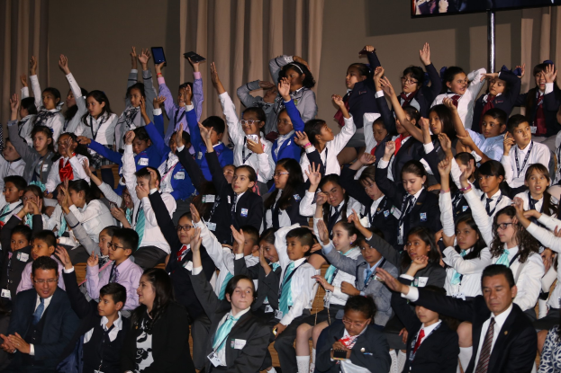 Gruppe von Kindern auf einer Bühne sitzend mit einem Mann und einer Frau davor, Bühne dekoriert mit Vorhängen und einer Wand, Fernseher auf einem Ständer im Hintergrund.
