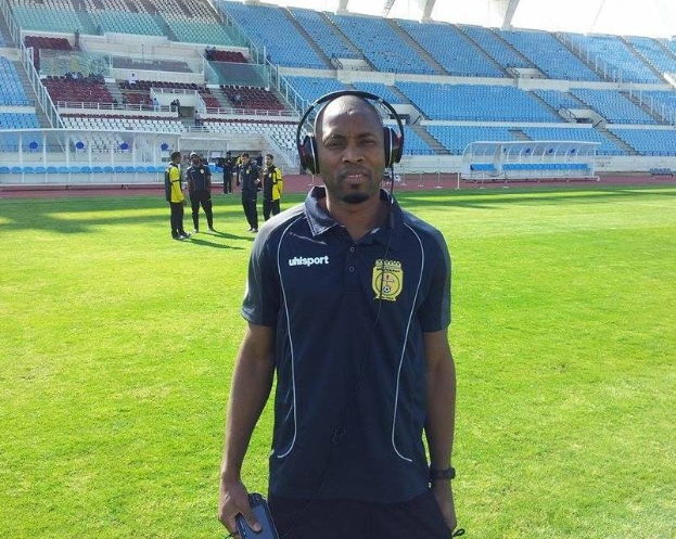 Ein Mann mit Headset, wahrscheinlich der Trainer der ghanaischen Fußballnationalmannschaft, steht auf einem Fußballfeld neben einem Stadion, hält ein Objekt und ist von ein paar Leuten im Hintergrund unter einem Himmel umgeben.