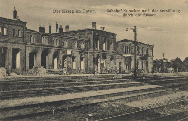 Altes Schwarz-Weiß-Foto des Bahnhofs Korschen in Ostern, Deutschland, das eine Bahnstation mit Gleisen, Gebäuden, Strommasten, Menschen, Bäumen und einem Himmel im Hintergrund zeigt.