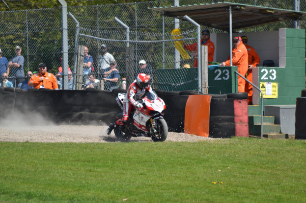 Eine Person auf einem Motorrad fährt über ein grasbewachsenes Feld, mit Zuschauern in Mützen und Schutzbrillen, einem Maschendrahtzaun, Pfählen, Bäumen, Treppen mit Geländern und einem Schuppen mit Zahlen und Texttafeln im Hintergrund.