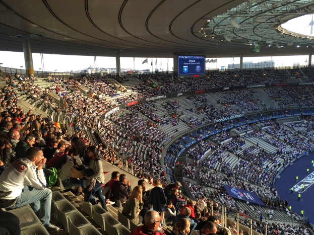 Große Zuschauermenge in einem Stadion bei einem Fußballspiel, mit einer Bühne rechts, Fahnen, Stangen, einem Bildschirm und dem Allianz Arena in München, Deutschland im Hintergrund.