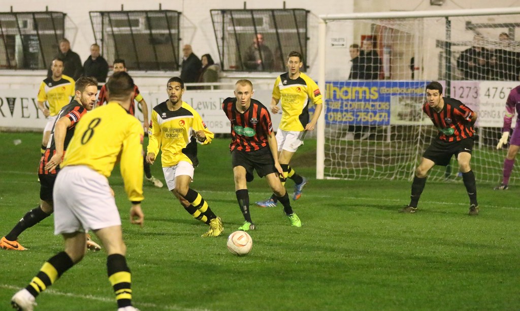 Ein Fußballfeld mit mehreren spielenden Männern, ein Netz auf der rechten Seite, eine Tafel mit Text im Hintergrund, Zuschauer hinter der Tafel und eine Wand mit Fenstern weiter hinten.