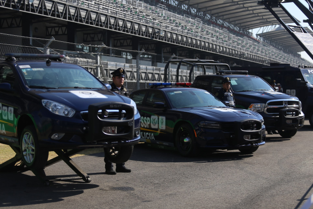 Eine Gruppe von Polizeiwagen vor einer Rennstrecke geparkt, mit uniformierten Personen auf der Straße, mit einem Stadion und Maschendraht im Hintergrund.