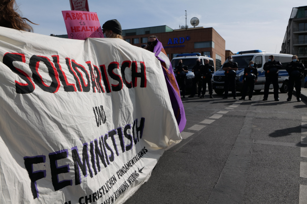 Eine Gruppe von Menschen marschiert auf einer Straße, hält ein Banner mit der Aufschrift "Solidarität und Feminismus" und fährt parkende Fahrzeuge und Gebäude im Hintergrund unter einem klaren blauen Himmel.