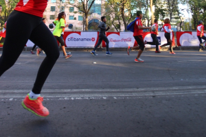 Eine Gruppe von Menschen, die einen Marathon auf einer Straße umgeben von Bäumen, Gebäuden mit Fenstern, Bannern mit Text, Laternen und einem klaren blauen Himmel laufen, einige tragen Rucksäcke und Turnschuhe.
