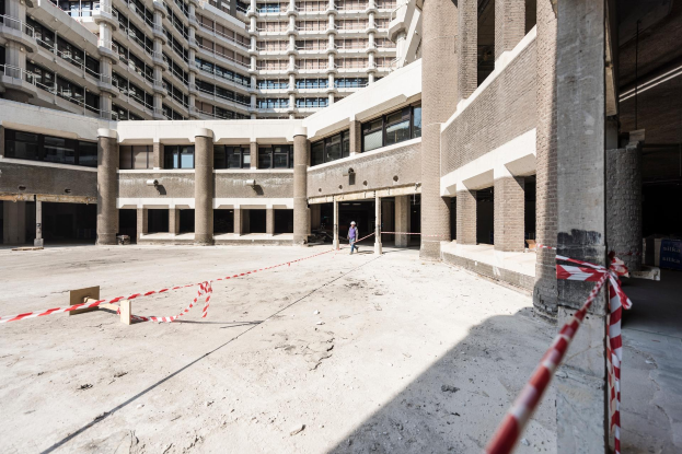 Großes Gebäude im Bau mit vielen Fenstern und Säulen, eine Person steht zentral und ein rotes und weißes Band ist an einem Pfahl auf der rechten Seite gebunden.