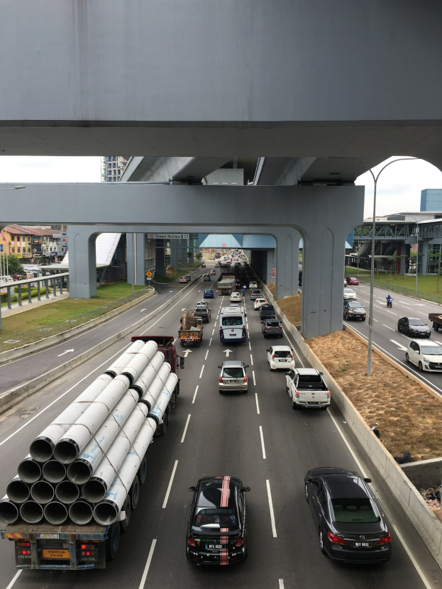 Luftaufnahme einer verstopften Autobahn unter einer Brücke, mit Fahrzeugen, Straßenlaternen, Strommasten, Grasflächen, Gebäuden und Bäumen im Hintergrund unter einem klaren Himmel.