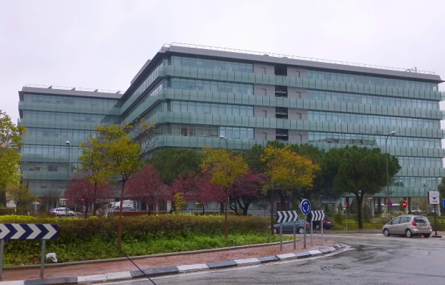 Großes modernes Gebäude mit vielen Fenstern, als Hauptquartier der Europäischen Zentralbank etikettiert, umgeben von Grünflächen, Straßeninfrastruktur und Fahrzeugen unter einem bewölkten Himmel.