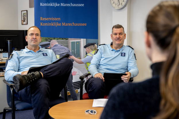 Zwei Polizeibeamte sitzen an einem Tisch mit Papieren, unterhalten sich mit einer Frau, während ein Monitor, eine Tastatur und andere Gegenstände auf einem nahen Tisch stehen, mit Fotorahmen und einer Uhr an der Wand.