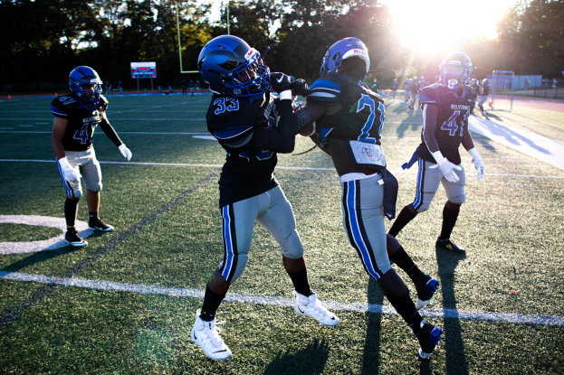 Gruppe junger Männer in Helmen und Schuhen beim Fußballspielen auf einem von Bäumen umgebenen Feld unter einem klaren blauen Himmel.