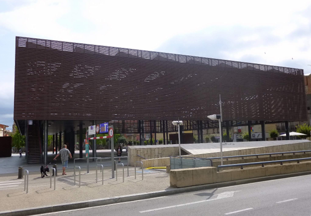 Großes modernes Gebäude mit zahlreichen Fenstern, das als neuer Eingang zur Stadtmitte dient, umgeben von städtischer Infrastruktur einschließlich Straßenlaternen, Fahrzeugen, Fußgängern und Bäumen unter einem bewölkten Himmel.