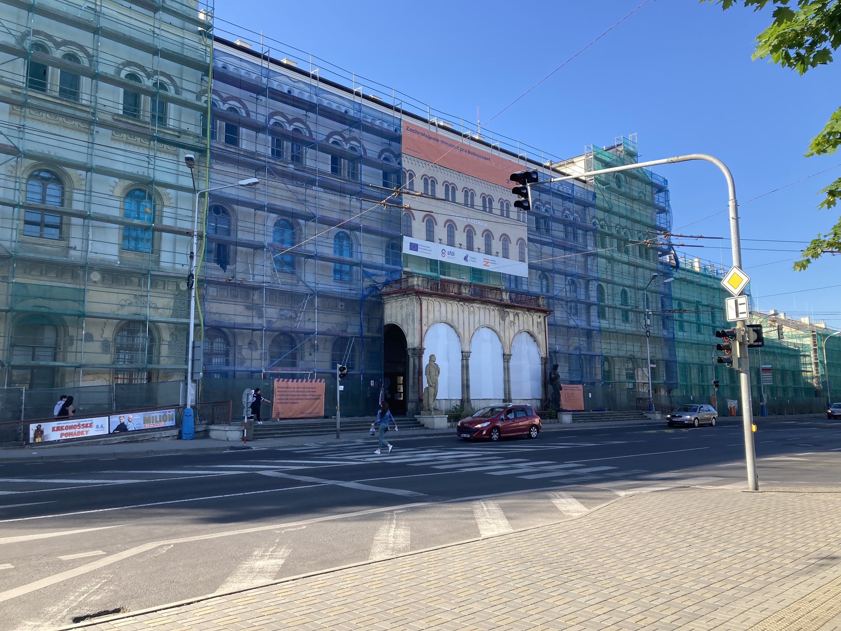 Ein großes Gebäude mit Gerüst, das renoviert wird, umgeben von Fahrzeugen, Fußgängern, Ampeln, Straßenschildern, Polen, Bäumen und einem klaren blauen Himmel.