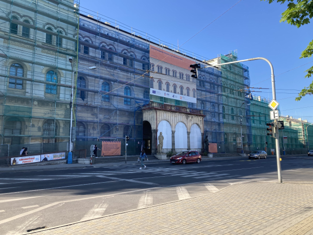 Ein großes Gebäude mit Gerüst, das renoviert wird, umgeben von Fahrzeugen, Fußgängern, Ampeln, Straßenschildern, Polen, Bäumen und einem klaren blauen Himmel.